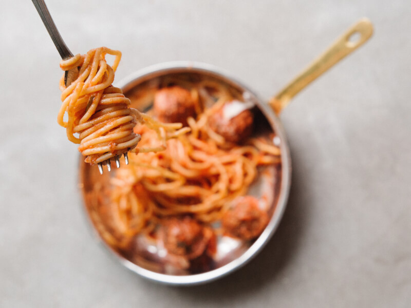 Close up of spaghetti in tomato sauce on a fork, with a pan full of pasta underneath, slightly blurred