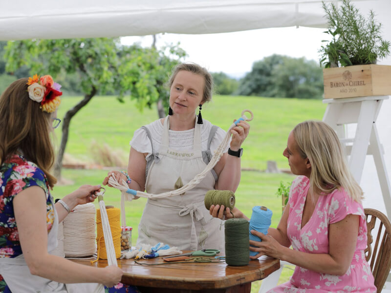 A woman is teaching two other women how to make a macrame plant holder in a large marquee