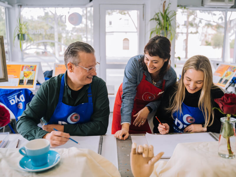 A man and woman are being taught to draw by a woman who is standing behind them