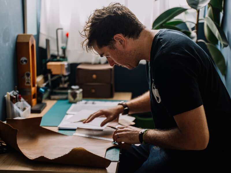 A man is sitting at a table, cutting pieces out of brown leather by hand using a scalpel