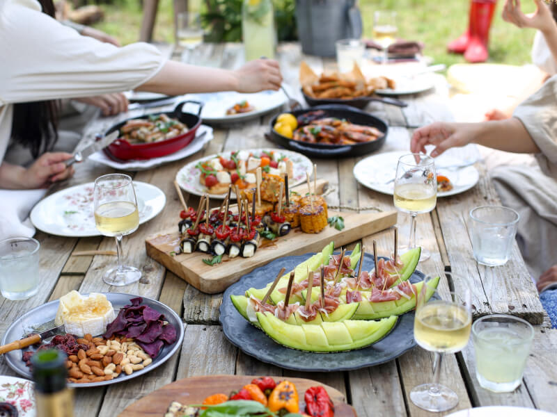 A selection of picnic foods sits on a wooden table, with hands selecting items