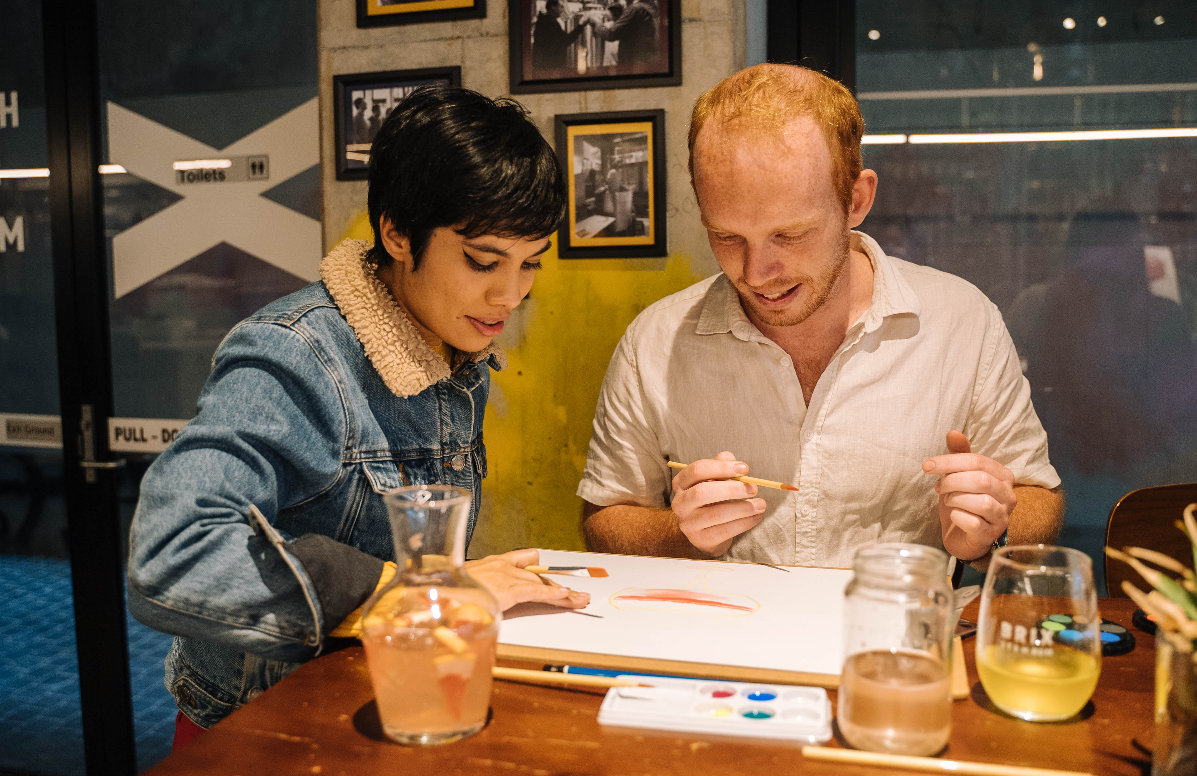A man and woman are sitting in a bar with drinks, at a paint and sip class