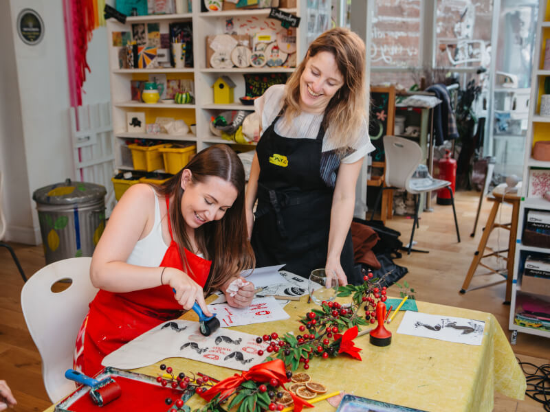 A person at a table is making a lino print Christmas stocking, while the craft teacher looks on