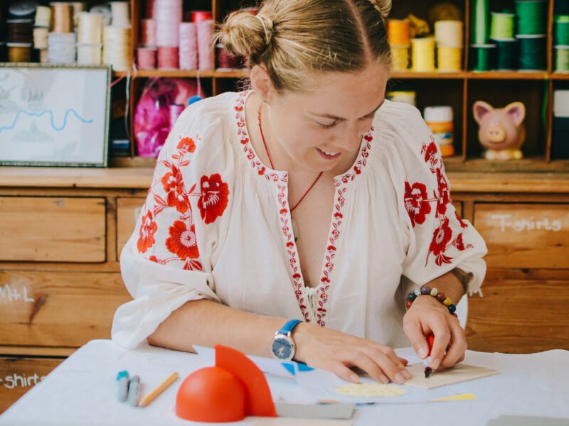 A woman is sitting at a table, carving a design into a block of lino