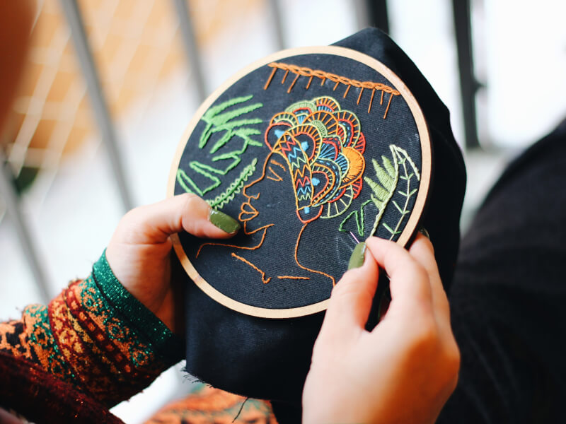 A person is sitting down, stitching a brightly coloured design onto an embroidery hoop
