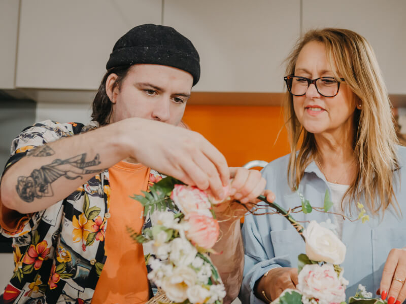 A man and woman sit at a table making a flower wreath using flowers and foliage