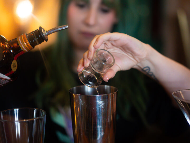A close up of a woman with green hair adding a measure of spirit to a cocktail shaker