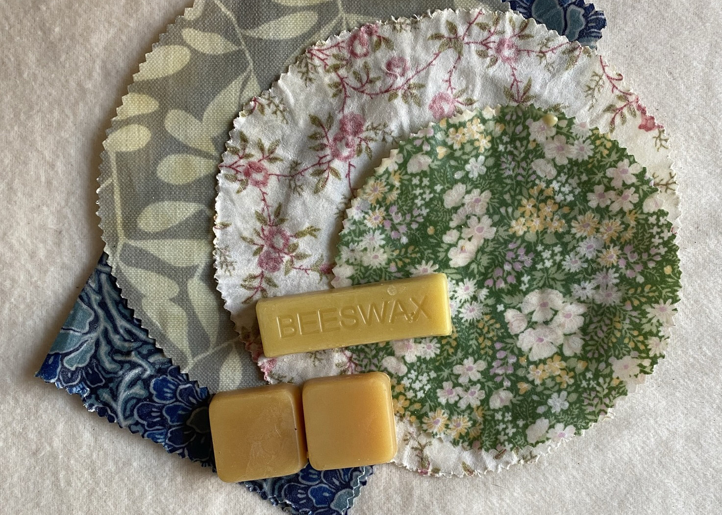 Handmade beeswax wraps in floral print sit on a table with beeswax blocks on top
