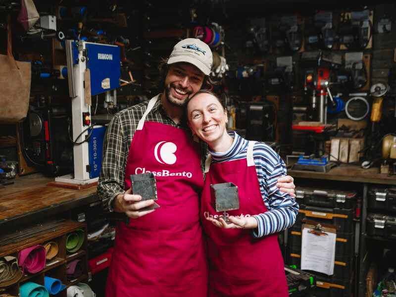 A man and woman stand cuddling, holding metal projects they made in a workshop