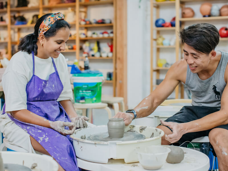 A man and woman sit in front of pottery wheels with freshly made pots on them