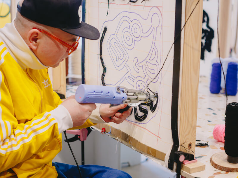 A man in a yellow jacket is creating a tufted rug with a tufting gun