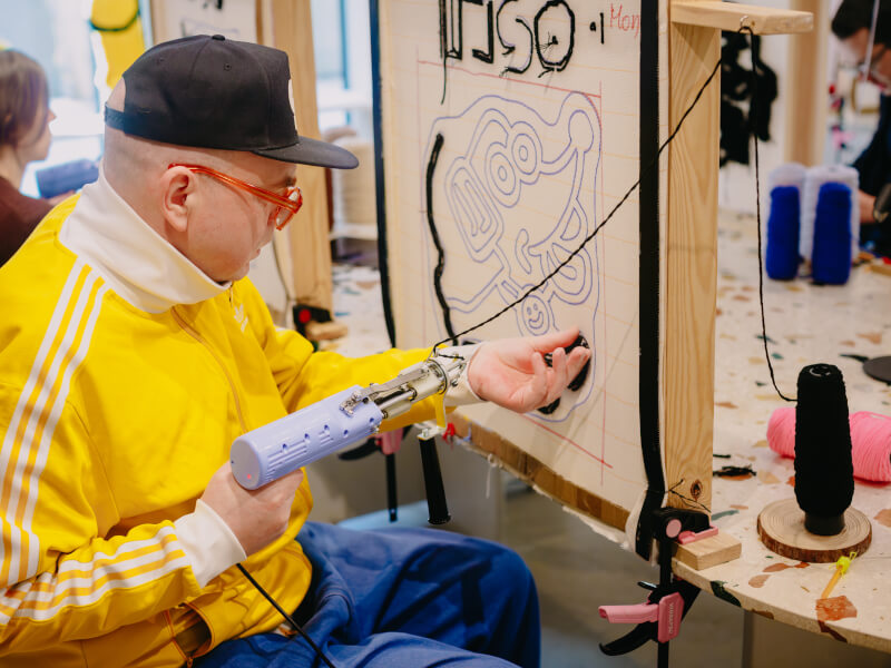 A man in a yellow tracksuit top is sitting at a canvas using a tufting gun