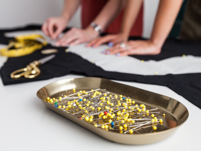 A tray of sewing pins sit on a table, there are two people in the background laying out fabric