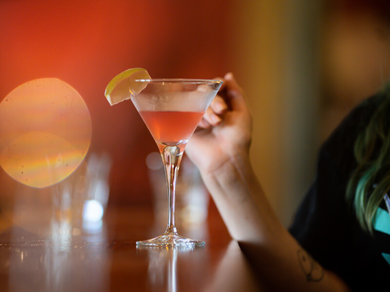 A red cocktail in a martini glass with a slice of lemon sits on a bar 