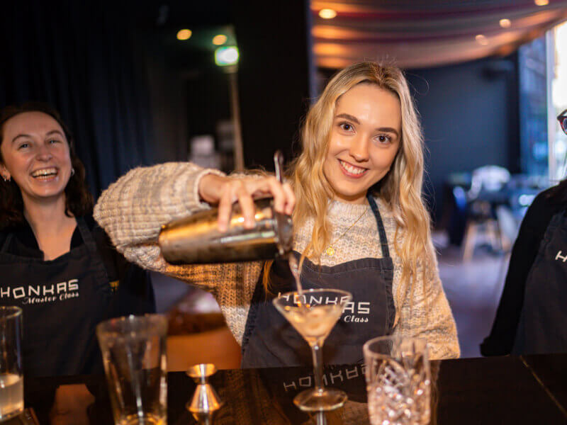 A woman is pouring a cocktail from a shaker into a glass and smiling at the camera