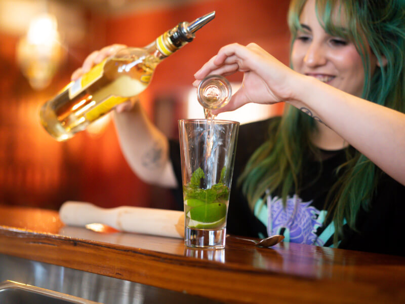 A woman with green hair is adding a shot measure of spirits to a glass