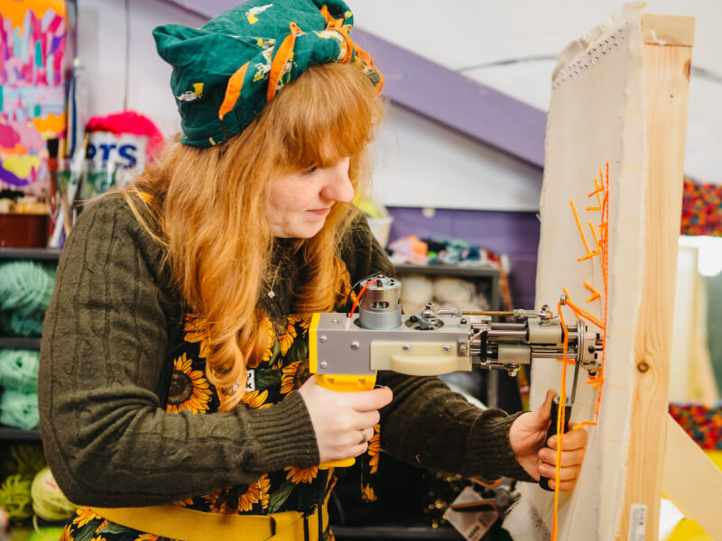 A woman is standing at a canvas with a tufting gun, creating a hand tufted design in orange wool.