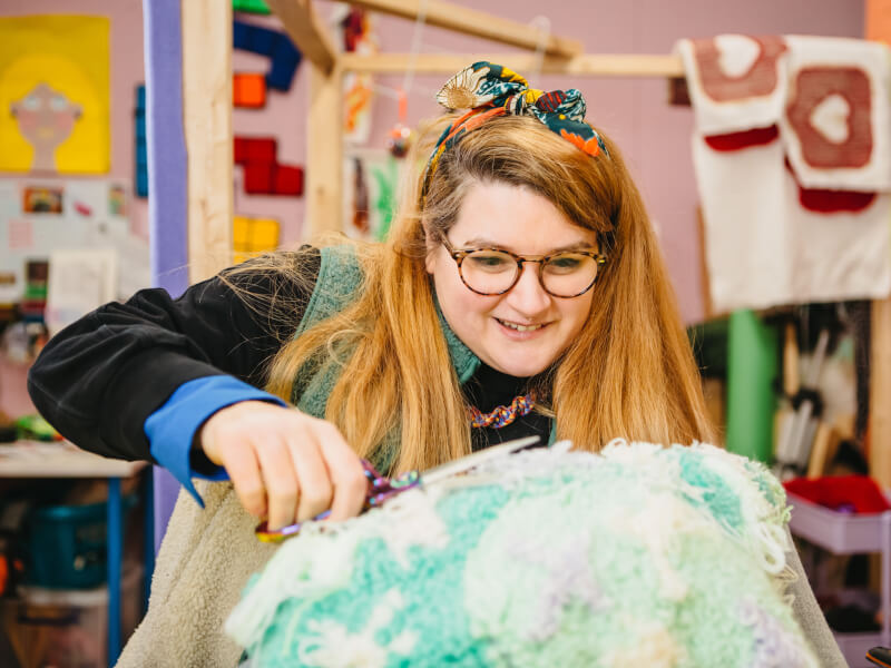 A woman is holding an aqua and white tufted project and is trimming the yarn to make it even all over.