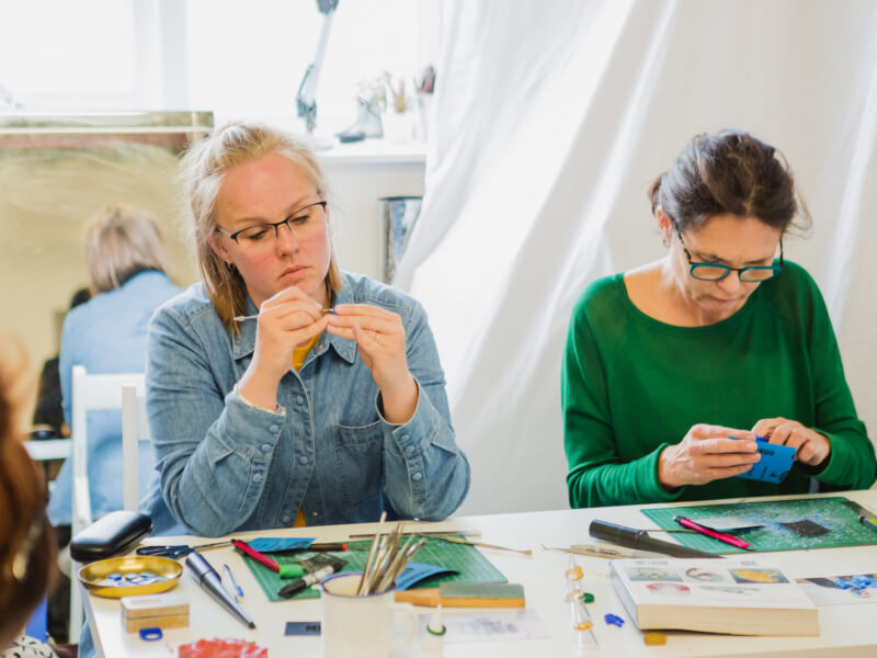 Two women sit at a table covered in jewellery making equipment, carving wax moulds 