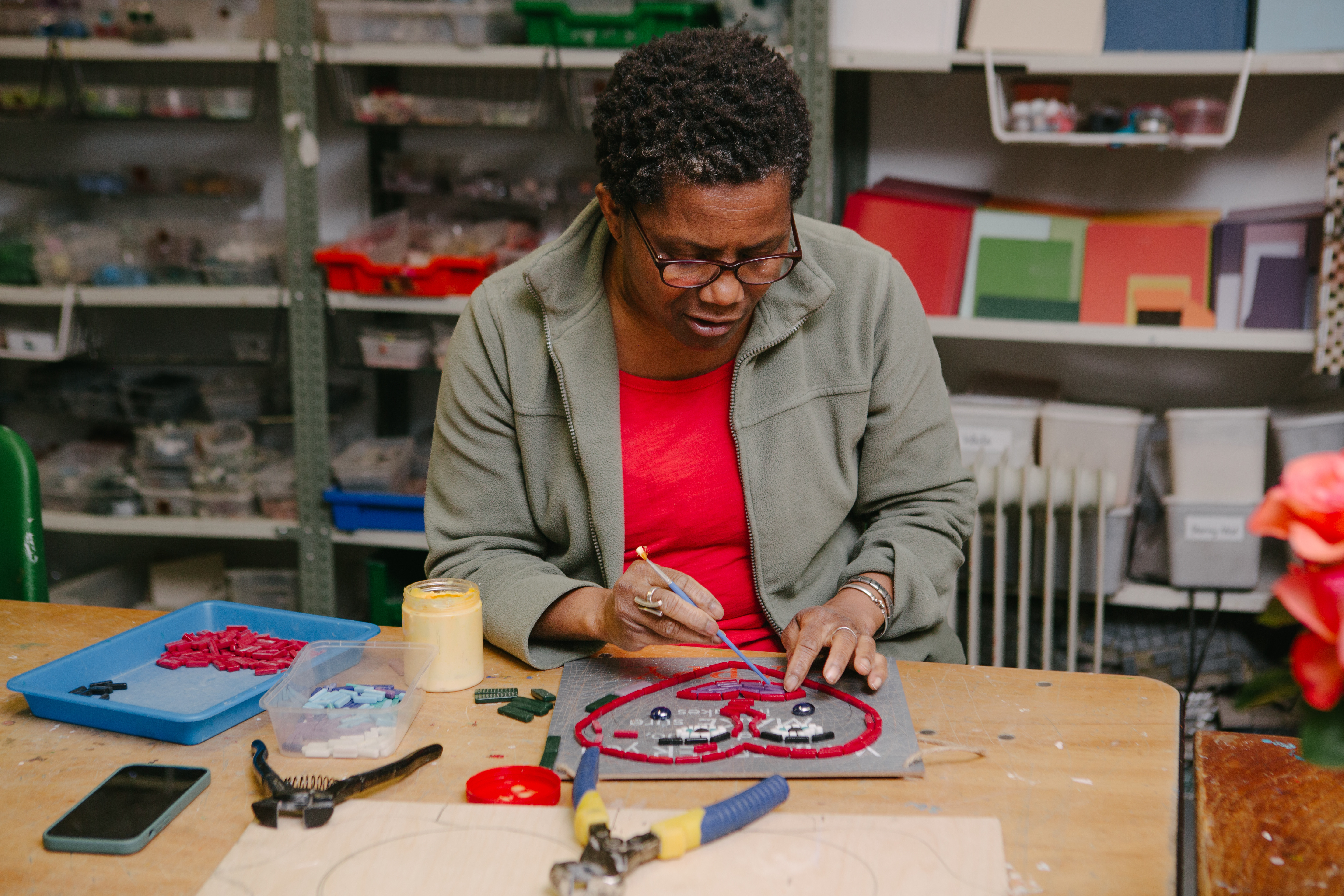 A woman is sitting at a table, making a piece of mosaic art from coloured tiles
