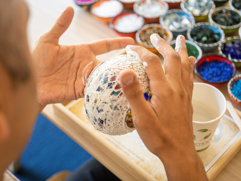 A close up of a person adding white grout to a coloured mosaic design on a lamp