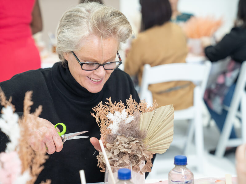 A woman is sitting at a table, creating a dried flower arrangement
