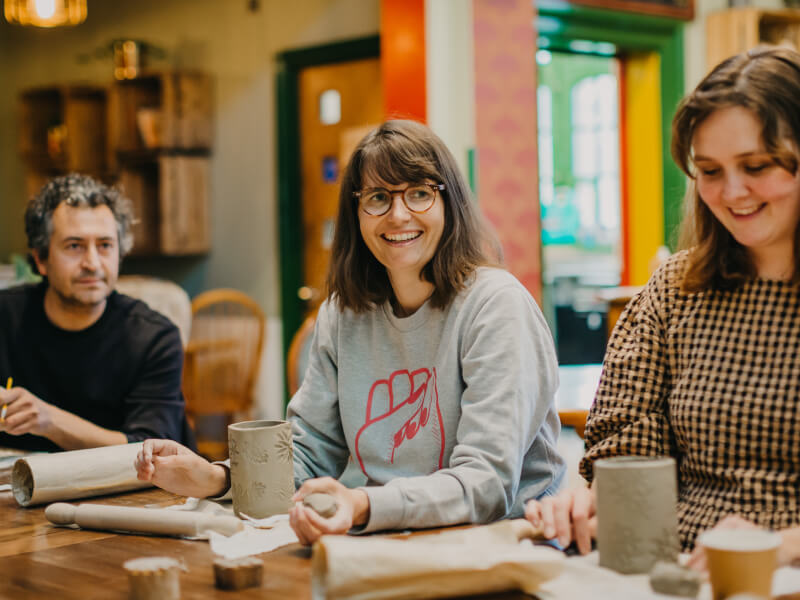 Three people are sitting at a wooden table, making handbuilt clay pots and laughing