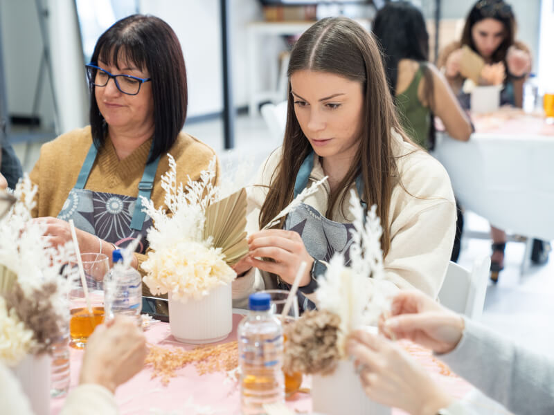 Two women are sitting at a table, making dried flower arrangements
