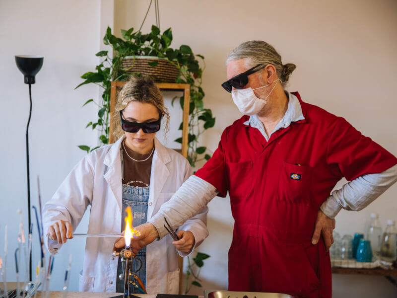 A man is teaching a woman in a white lab coat how to melt glass to blow