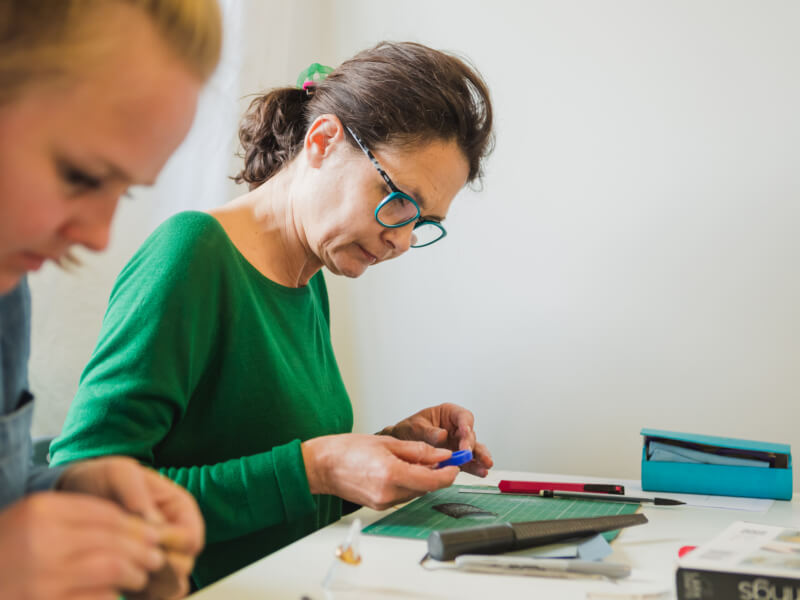 Two women are sitting at a table, sanding plastic templates in a jewellery making class