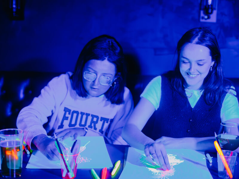 Two people are sitting at a table drawing at a neon life drawing class