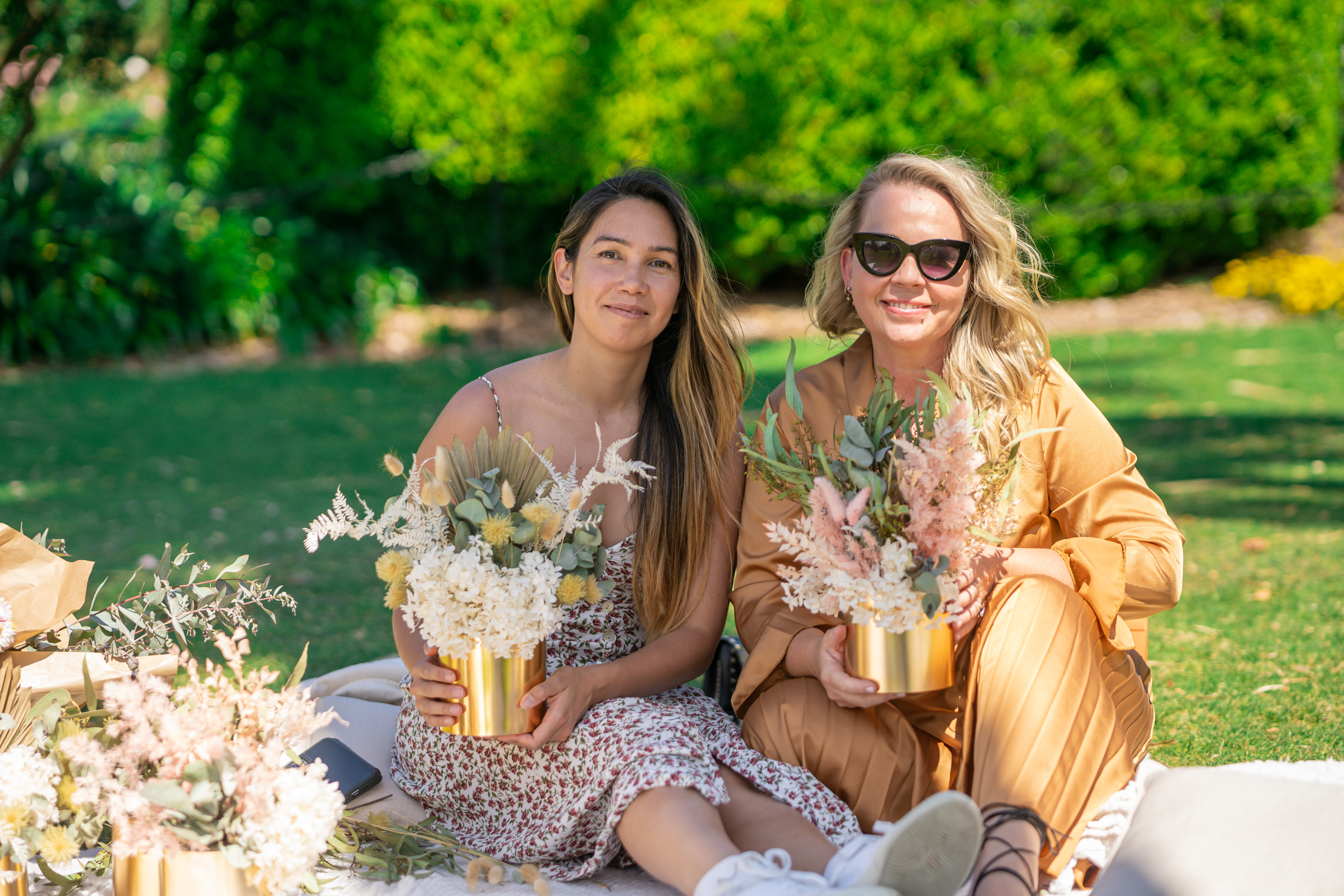 Two women are holding flower arrangements they've made at an outdoor class