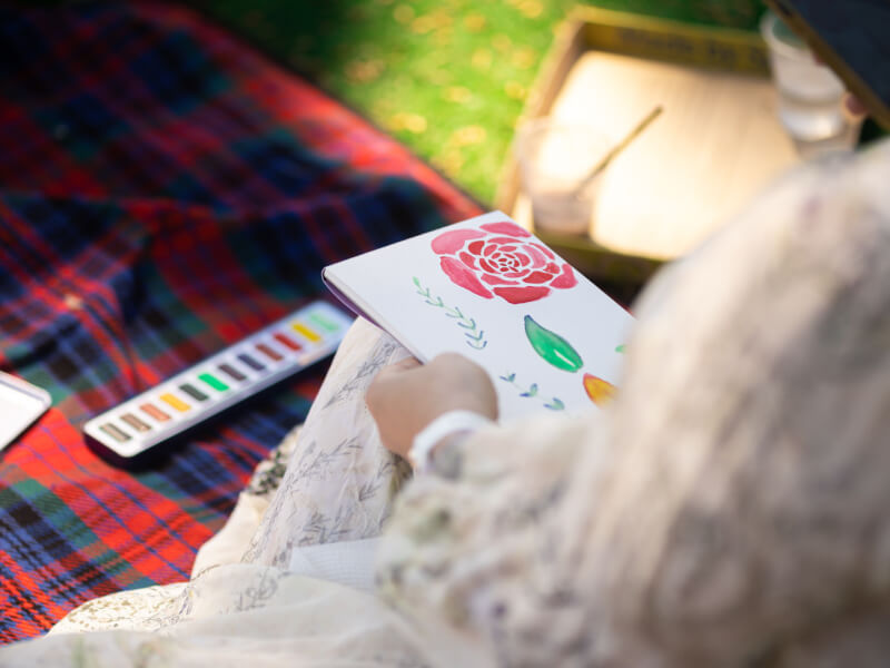 A close up of a person sitting on a rug, painting with watercolours outdoors