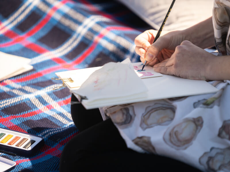 A close up picture of a person painting a watercolour, sitting outside on a blanket