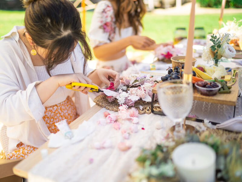 Two women are sitting at a table outside, making floral wreaths
