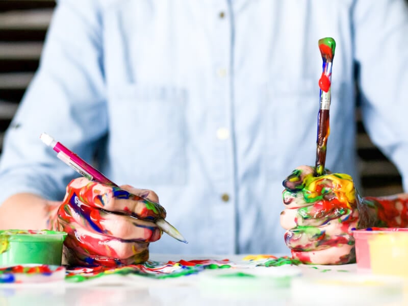 A man grips a paint brush in his hand and is covered in multicoloured paint
