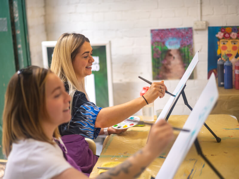 Two women are sitting at easels, learning to paint at a paint and sip class