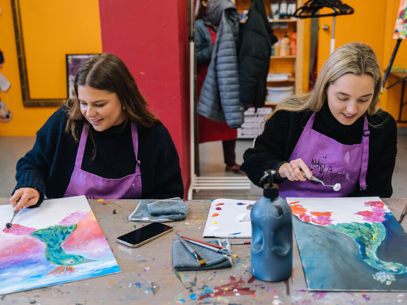 Two women are sitting at a table, painting colourful pictures of peacocks