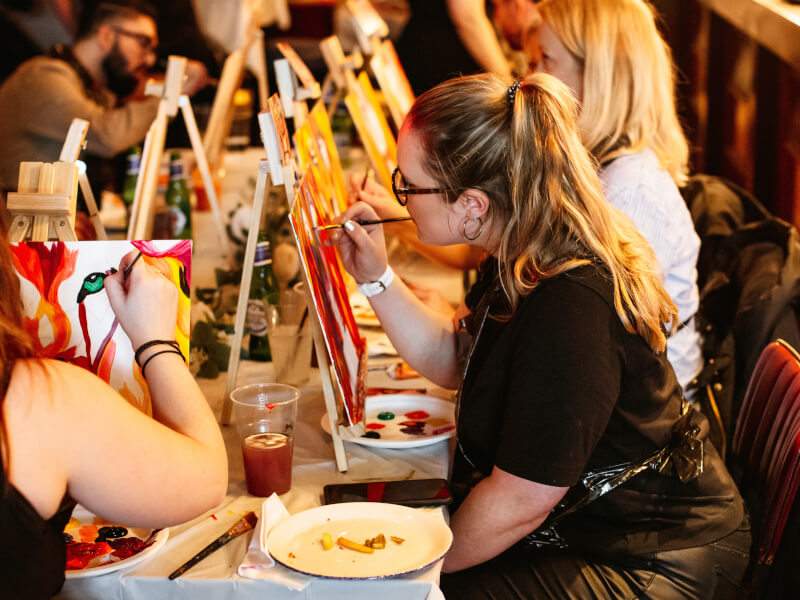 A group of people at a paint and sip class, painting pictures of lions in red and orange paint