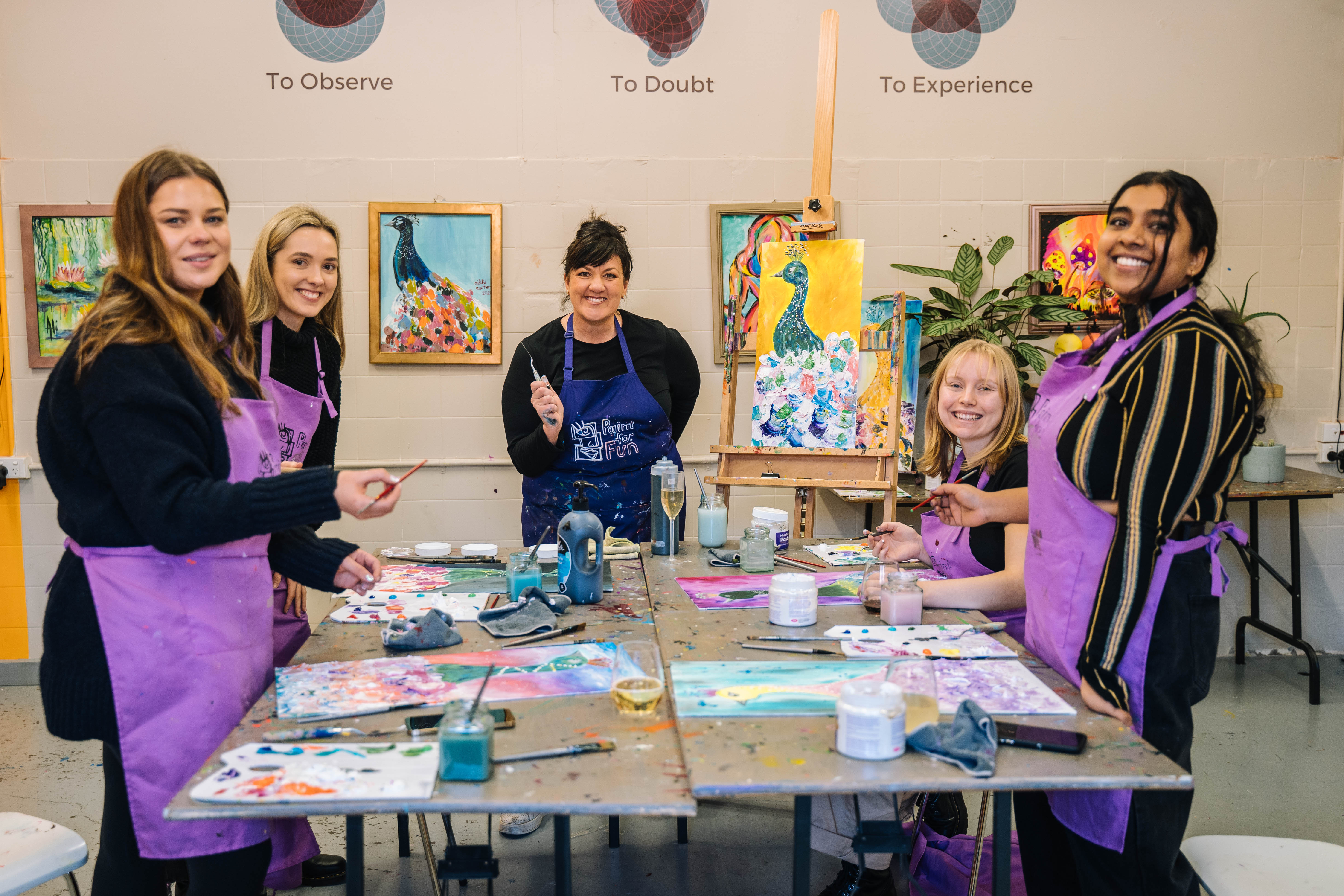 A group of women in purple aprons are standing around a table, surrounded by painting equipment