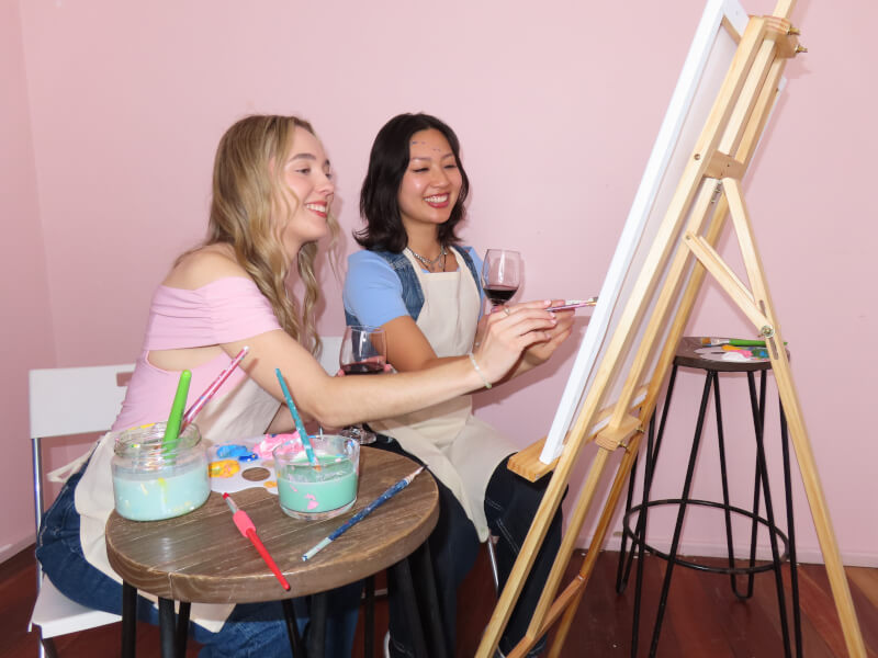 Two women smiling and painting on a canvas in a painting class