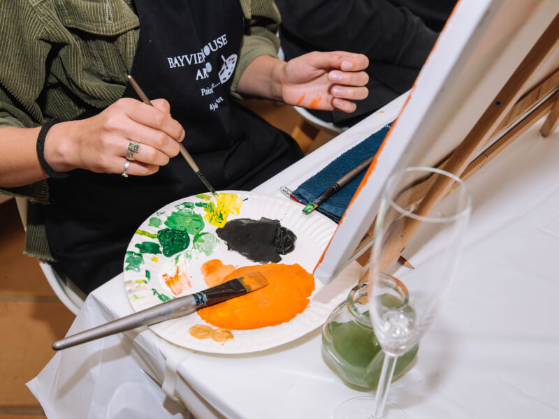A woman dips her paint brush into paint on a messy pallet at a painting class.