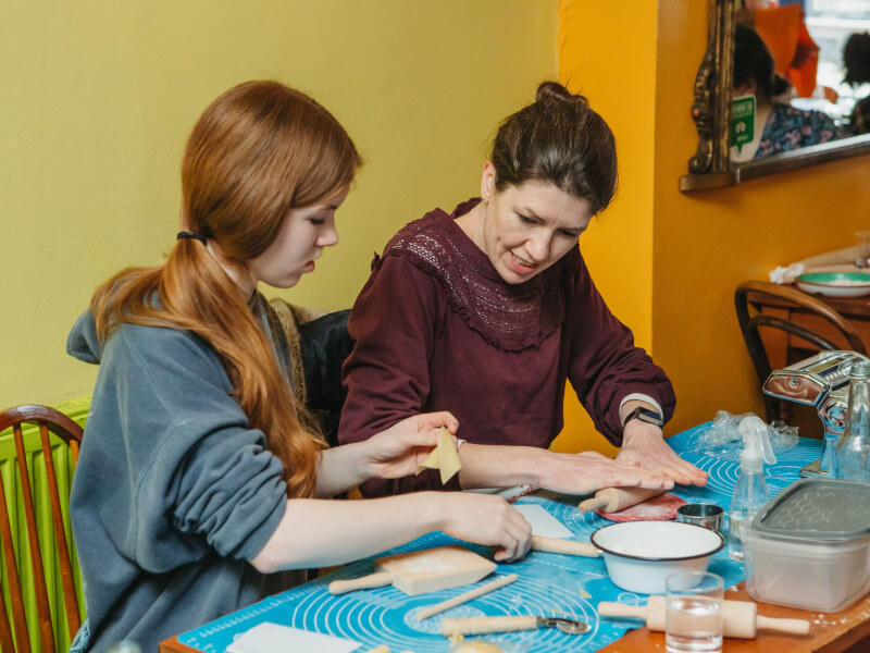 Two people sitting at a table making pasta shapes from red dough