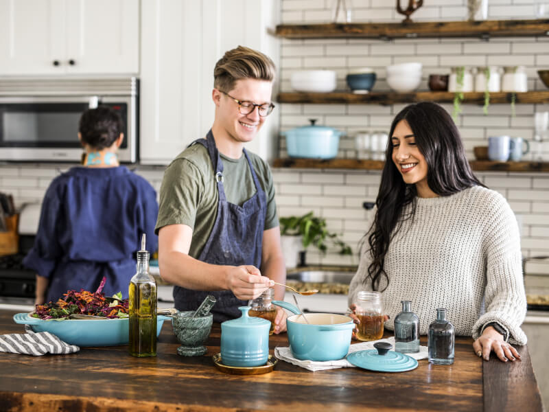 Three people are in a kitchen cooking. There are a variety of oils and a large bowl of salad on the table