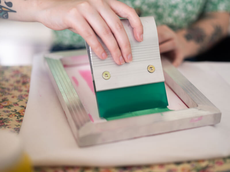A close up of a person drawing a blade over green ink during the screenprinting process