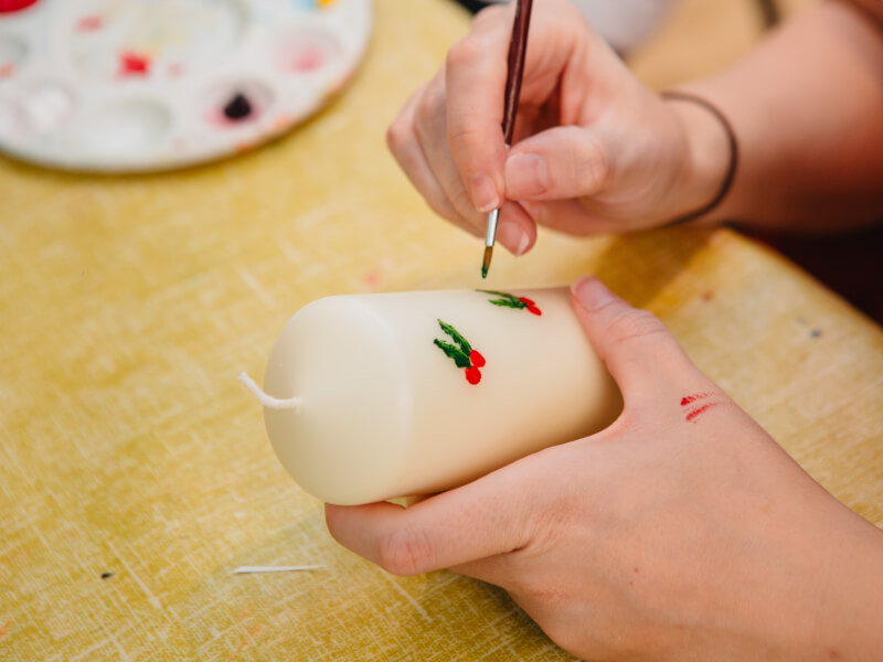 A close up of a person painting holly berries and leaves on a white pillar candle