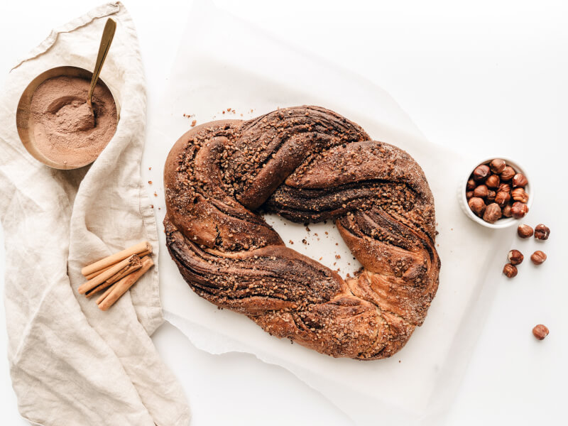 A circular twisted loaf of bread with a cinnamon stick and a small bowl full of chocolate powder sit on a cream tablecloth