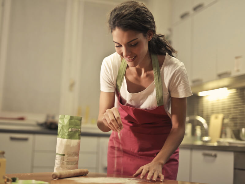 Woman wearing red apron smiling whilst sprinkling flour. 