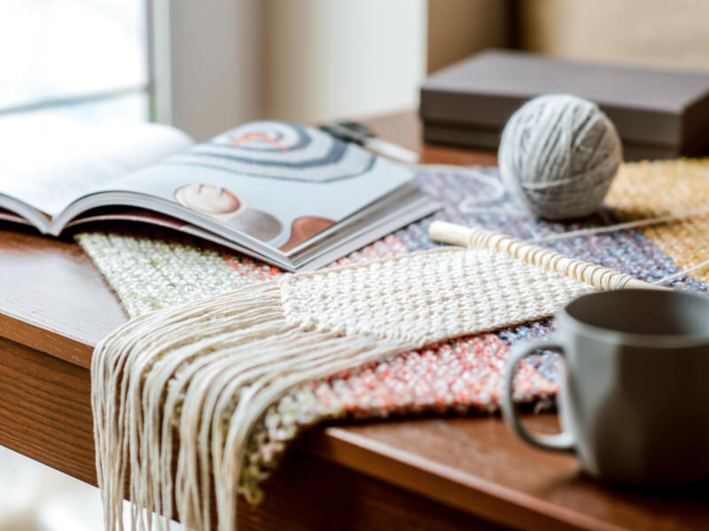 A macrame wallhanging lies on a table, there's a book, ball of yarn and cup beside it
