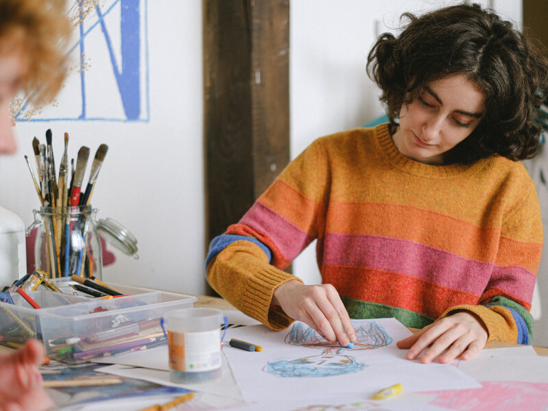 A woman in a stripy jumper sits at a table sketching in coloured chalks
